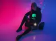 Woman in cyberpunk outfit sitting in neon-lit room, wearing a black hoodie with glowing green NEO logo – crypto streetwear aesthetic