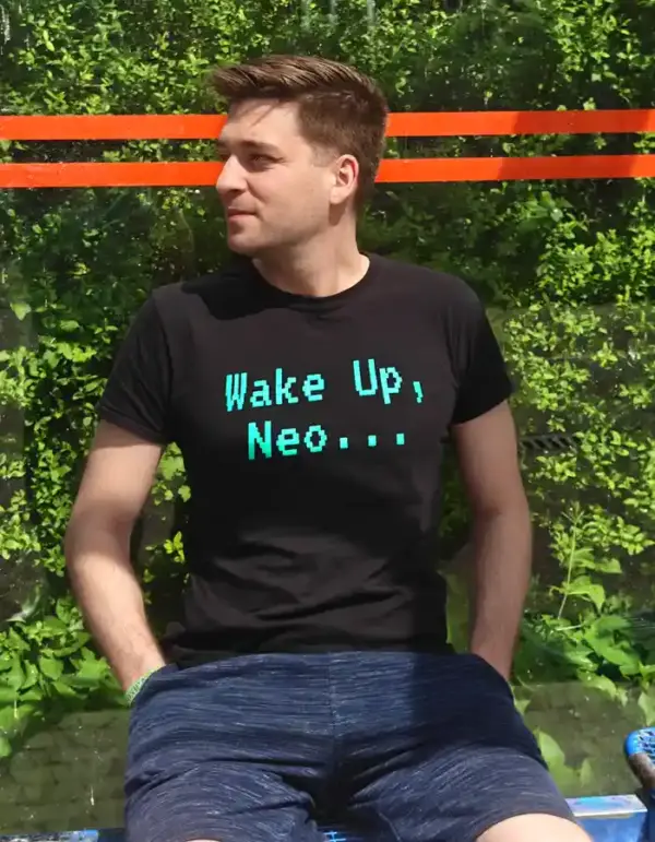 Man wearing a black "Wake Up, Neo..." T-shirt while sitting outdoors in front of a green urban background.