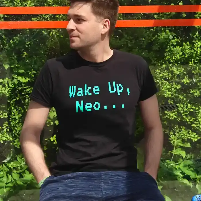 Man wearing a black "Wake Up, Neo..." T-shirt while sitting outdoors in front of a green urban background.